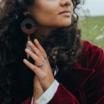 A woman with curly hair wearing vintage earrings and rings, standing in a grassy meadow.