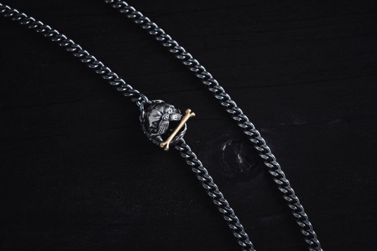 Close-up of a silver skull chain necklace with gold crossbones charm on a dark wooden background.